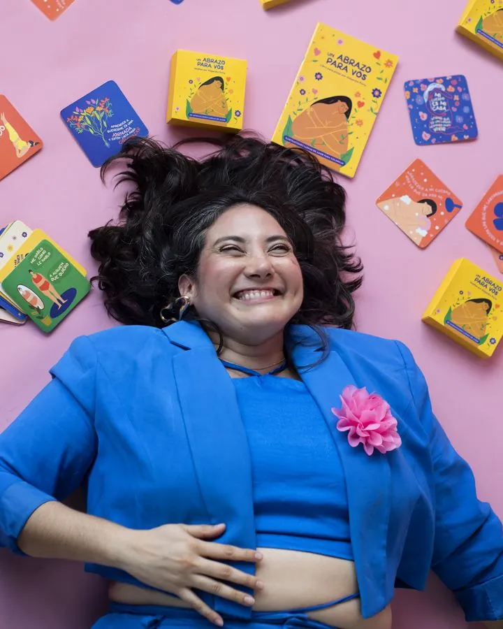 Ale laughing on a pink background, surrounded by her illustrated affirmation cards and colouring book